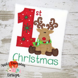 May include: Red and green Christmas themed embroidered design on a white shirt. The design features a large number "1" with snowflakes and a reindeer with a green and red scarf. The text "1st Christmas" is embroidered below the design.