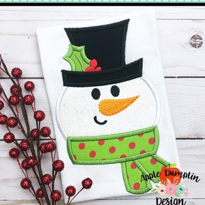 May include: A white snowman applique with a black top hat, green holly, red berries, a green scarf with red polka dots, and an orange nose. The applique is on a white shirt with the text "Apple Dumplin Design" in the bottom right corner.