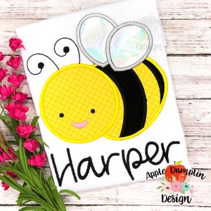 May include: A white t-shirt with a yellow and black embroidered bee with iridescent wings. The name "Harper" is embroidered below the bee in black cursive font. The text "Apple Dumplin Design" is also embroidered in black cursive font below the name.