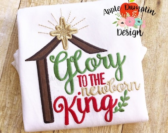 May include: A white t-shirt with an embroidered design of a manger with a star, the words "Glory to the Newborn King" and a small green sprig. The design is in red, green, brown, and gold thread.
