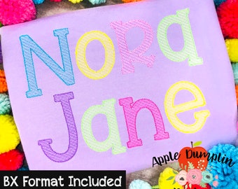 May include: A lavender-colored shirt with the name "Nora Jane" embroidered in colorful thread. The letters are in shades of blue, yellow, pink, and green. The text "BX Format Included" is at the bottom left. The design is surrounded by colorful pom-poms.