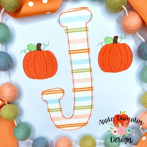 May include: A light blue fabric with a striped letter J and two orange embroidered pumpkins. The text "Apple Dumplin Design" is in the bottom right corner.