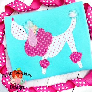 Può includere: Una maglietta blu turchese con un'applicazione rosa e bianca di un barboncino. Il barboncino ha un fiocco rosa e pois rosa. La maglietta è decorata con un nastro rosa con pois bianchi.