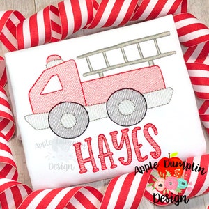 May include: White t-shirt with a red and gray embroidered fire truck design. The fire truck has a ladder, wheels, and a siren. The name "HAYES" is embroidered in red below the truck. Red and white striped ribbon is in the background.
