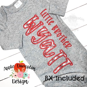 May include: A gray baby onesie with long sleeves. The onesie features the embroidered text "Little Brother Wyatt" in red thread with a plaid pattern. The design includes the text "BX Included" in black.
