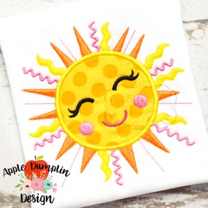May include: Embroidered design of a smiling sun with yellow polka dots and pink cheeks. The sun has orange rays and pink and yellow wavy lines around it.