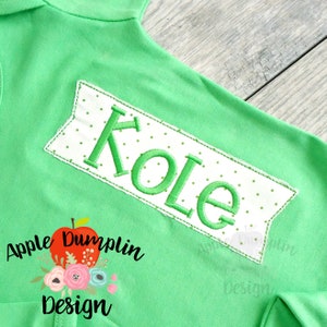 May include: Green and white banner with the name "Kole" embroidered in green thread. The banner has a white background with green polka dots. The banner is sewn onto a green shirt. The shirt has a small apple and flower design on the bottom left corner with the text "Apple Dumplin Design".