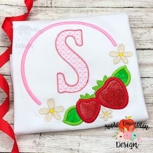 May include: A white t-shirt with a pink circle and the letter "S" inside. The circle is surrounded by two strawberries, two white flowers, and green leaves. The text "Apple Dumplin Design" is at the bottom of the shirt.