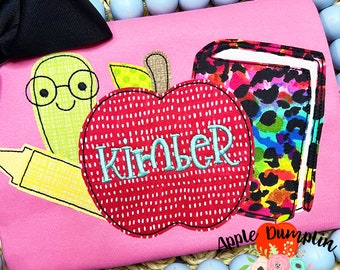 May include: Pink shirt with an embroidered apple, a book, and a crayon. The apple has the name "Kimber" embroidered on it. The book has a colorful, animal print design. The shirt has the text "Apple Dumplin Design" at the bottom.