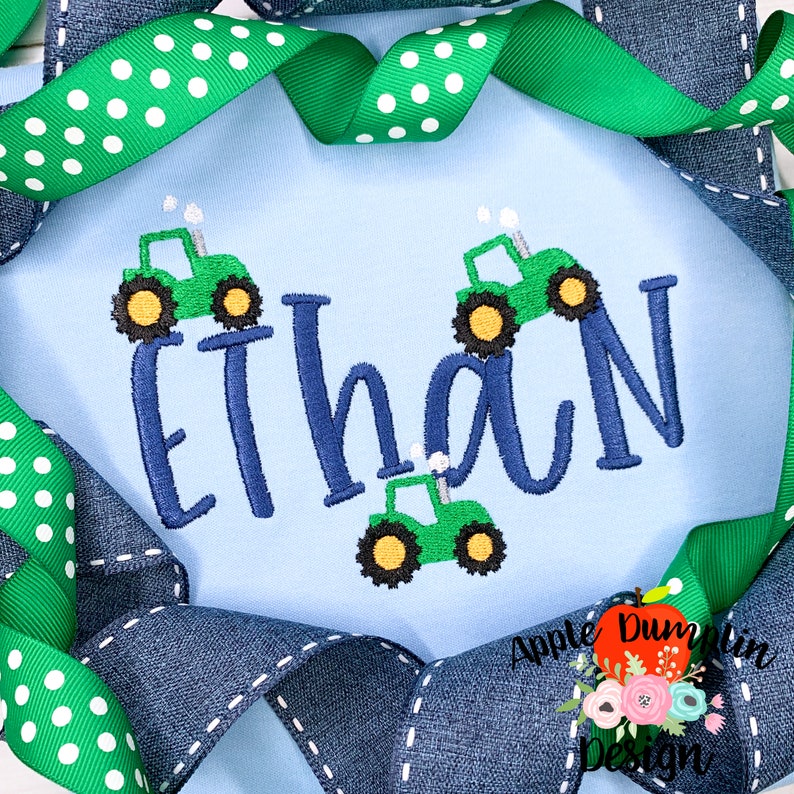 May include: A light blue baby onesie with the name "Ethan" embroidered in blue. Three green toy tractors with black wheels are embroidered on the onesie.