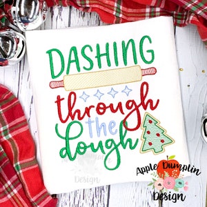 May include: Embroidered design on a white fabric with the text "Dashing through the dough" in green, red, and blue lettering. A rolling pin and a Christmas tree are also embroidered on the fabric.