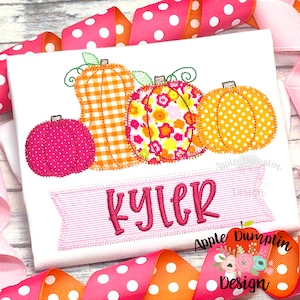 May include: White t-shirt featuring embroidered pumpkins in various patterns and colors, including orange gingham, floral, and polka dots. The name "Kyler" is embroidered on a pink banner. Ribbons in pink and orange with white polka dots frame the shirt.