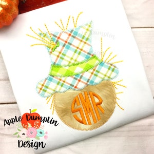 May include: A brown and tan scarecrow applique with a plaid hat and a monogrammed circle. The monogram is the letters "SMR" in orange thread. The scarecrow is surrounded by yellow stitching that looks like straw.