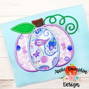 May include: A light blue t-shirt with a purple, blue, and pink paisley patterned pumpkin applique. The pumpkin has a green stem and brown stem. The text "Apple Dumplin Design" is in the lower right corner of the image.
