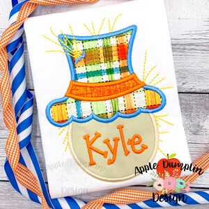 May include: A white t-shirt with an embroidered applique of a scarecrow head with a plaid hat and the name "Kyle" on a circle. The scarecrow has a yellow straw-like hair and a blue and orange hat.