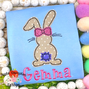 May include: A light blue t-shirt with a brown bunny with pink polka dots and a pink bow. The bunny has a purple flower on its back. The name "Gemma" is embroidered in pink below the bunny.