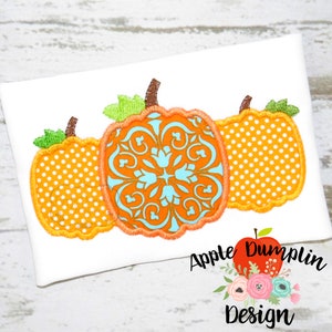 May include: Three embroidered pumpkins in orange, white and blue. The centre pumpkin has a blue and white floral pattern. The pumpkins are on a white background with the text "Apple Dumplin Design".