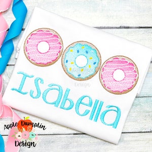 May include: A white t-shirt with a blue and pink donut design and the name "Isabella" embroidered in blue. The shirt is folded and has a pink and blue ribbon on the left side.