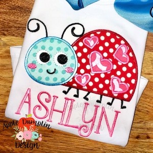 May include: A white t-shirt with a red and white ladybug with pink hearts and a pink embroidered name "ASHLYN".