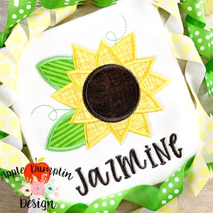 May include: A white onesie with an embroidered sunflower and the name "Jasmine" in black cursive lettering. The sunflower has a yellow center with brown petals and green leaves.