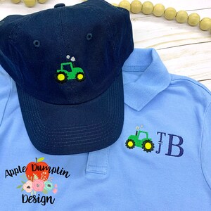 May include: A blue baseball cap with a green tractor embroidered on the front. A light blue polo shirt with a green tractor and the initials "TB" embroidered on the front. The shirt has a red apple with a green leaf and the text "Apple Dumplin Design" embroidered on the bottom left.
