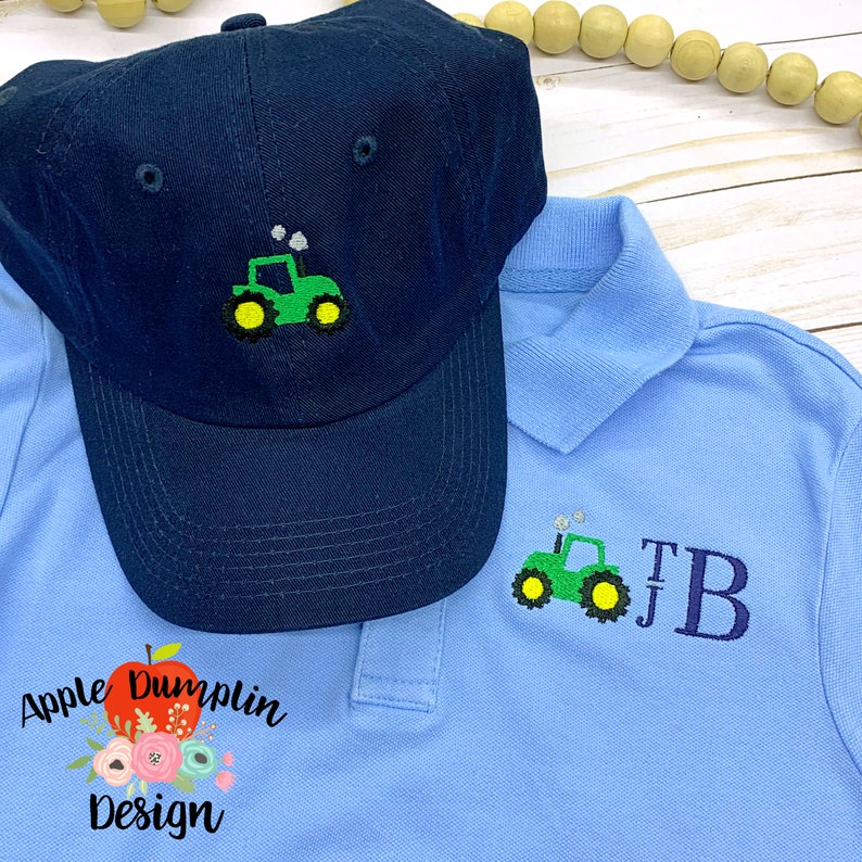 May include: A blue baseball cap with a green tractor embroidered on the front. A light blue polo shirt with a green tractor and the initials "TB" embroidered on the front. The shirt has a red apple with a green leaf and the text "Apple Dumplin Design" embroidered on the bottom left.