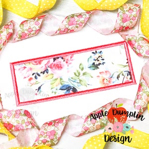 May include: A rectangular embroidery design featuring a floral pattern with pink, blue, and yellow flowers on a white background. The design is outlined with a red border.