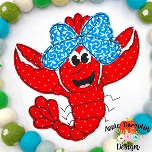 May include: Red crawfish with white polka dots wearing a blue bow with a white floral pattern. The crawfish is smiling with big eyes and has a red tail.