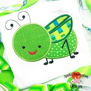 May include: Green embroidered applique of a cartoon grasshopper with a checkered wing, polka dot wing, and a smiling face.