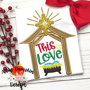 May include: Embroidered design of a manger scene with a star above it and the text "This is Love". The manger is brown, the star is gold, and the text is red, green, and blue.
