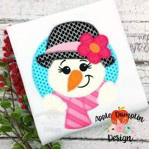Op de afbeelding: Een schattige sneeuwpop-applique met een roze-wit gestreepte sjaal, een zwart-witte stippenhoed en een roze bloem. De applique is op een witte achtergrond met de tekst "Apple Dumplin Design" in de rechterbenedenhoek.