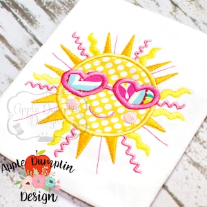 May include: Embroidered design of a yellow sun with a smiling face wearing pink heart-shaped sunglasses with white and blue accents. The sun has yellow rays and a white polka dot pattern.
