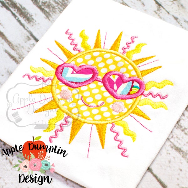 Pode incluir: Desenho bordado de um sol amarelo com um rosto sorridente que usa óculos de sol em forma de coração rosa com detalhes brancos e azuis. O sol tem raios amarelos e um padrão de bolinhas brancas.