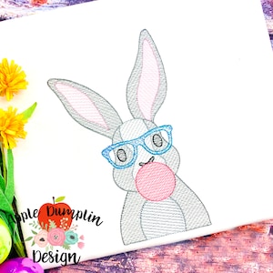 Può includere: Disegno ricamato di un coniglio grigio con occhiali blu e che tiene una palla rosa. Il coniglio guarda a destra. Il disegno è su uno sfondo bianco.