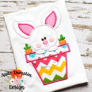 May include: Embroidered design of a white bunny peeking out of a pink and green flower pot with a chevron pattern. The bunny has pink ears and a brown nose.