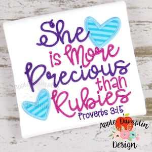 Op de afbeelding: Borduurwerk op een wit T-shirt met twee blauw-wit gestreepte harten. De tekst luidt "Zij is kostbaarder dan robijnen Spreuken 3:15".