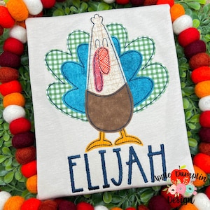 May include: A beige shirt with an embroidered turkey design. The turkey is blue, brown, and green with a red wattle. The shirt has the name "Elijah" embroidered in navy blue below the turkey.
