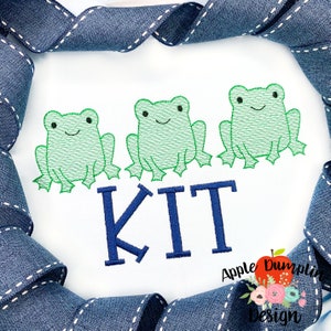 May include: Three green embroidered frogs sitting on a white background with the name "KIT" embroidered in blue below them.