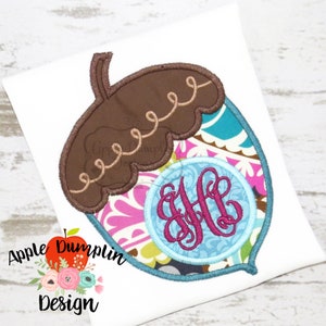 Könnte beinhalten: Eine braune und blaue Eichel mit einem Monogramm in einem Kreis. Das Monogramm ist in Pink und der Kreis ist auf einem blauen Hintergrund. Die Eichel ist auf einem weißen Hintergrund.