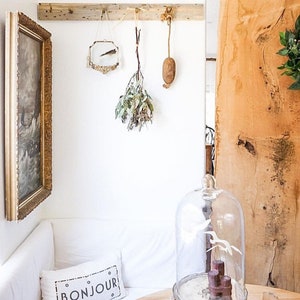 May include: A white couch with a pillow that says "BONJOUR" in black lettering. A glass cloche sits on a wooden table with a wooden tray and a candle. The wall behind the couch has a wooden shelf with a small mirror, a dried plant, and a brown object hanging from it.