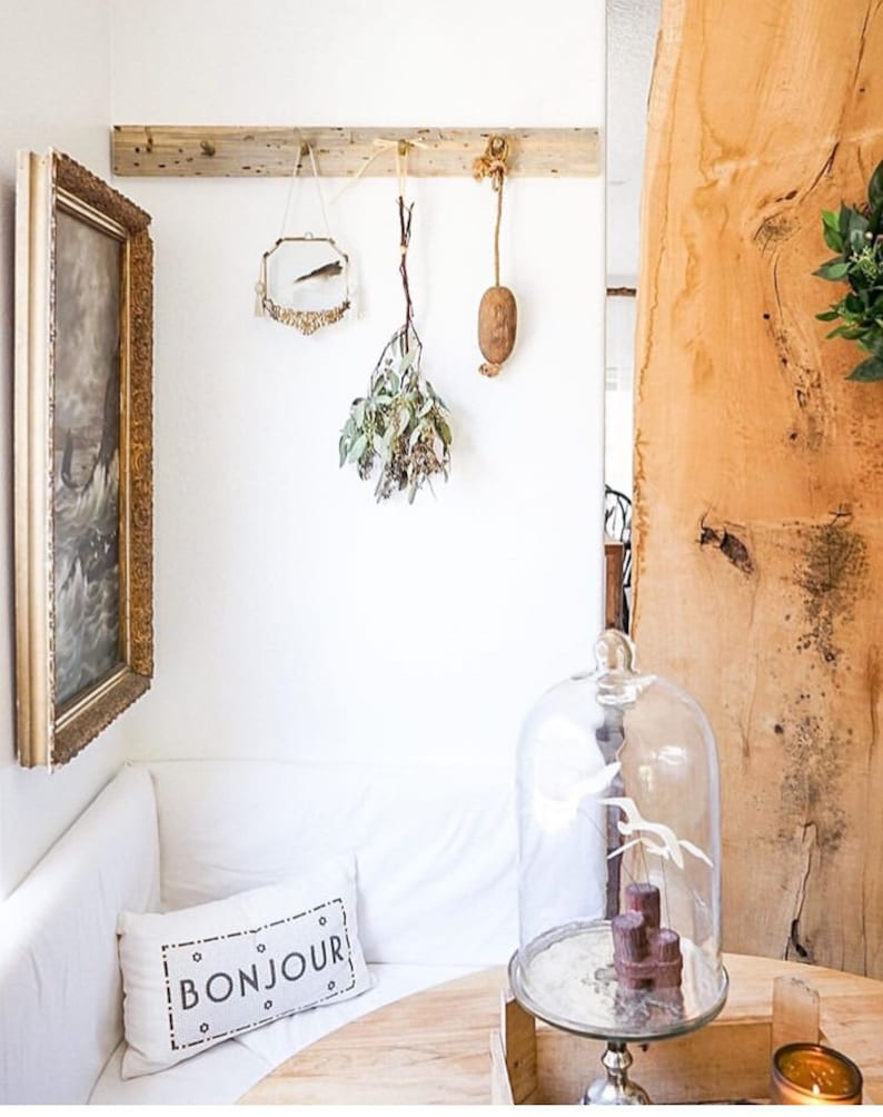 May include: A white couch with a pillow that says "BONJOUR" in black lettering. A glass cloche sits on a wooden table with a wooden tray and a candle. The wall behind the couch has a wooden shelf with a small mirror, a dried plant, and a brown object hanging from it.
