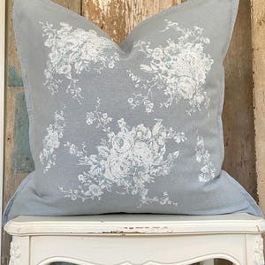 May include: A square, light blue pillow with a white floral pattern. The pillow features a detailed design of roses and other flowers. The pillow is resting on a white, ornate chair.
