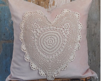 May include: A blush-colored square pillow with a large, cream-colored, heart-shaped crochet doily. The pillow is made of a soft, velvety material. The doily is centered on the pillow and has a detailed, circular design.