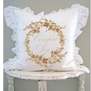 May include: A white decorative pillow with a ruffled edge. The pillow features a gold floral wreath design with the words "Comfort & Joy" in elegant script. The pillow is resting on a distressed white table.