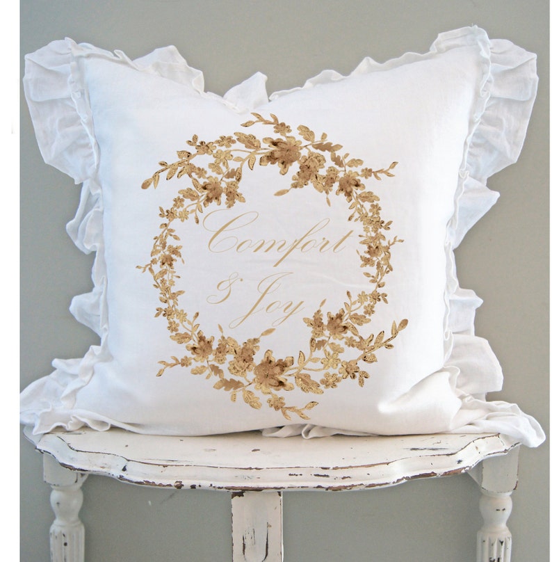 May include: A white decorative pillow with a ruffled edge. The pillow features a gold floral wreath design with the words "Comfort & Joy" in elegant script. The pillow is resting on a distressed white table.