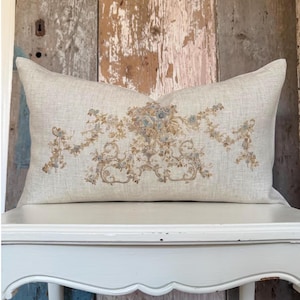 May include: A rectangular throw pillow made of light beige linen fabric. The center of the pillow features an intricate, symmetrical embroidered design in muted shades of dusty blue, sage green, and tan, depicting floral garlands and scrolls.