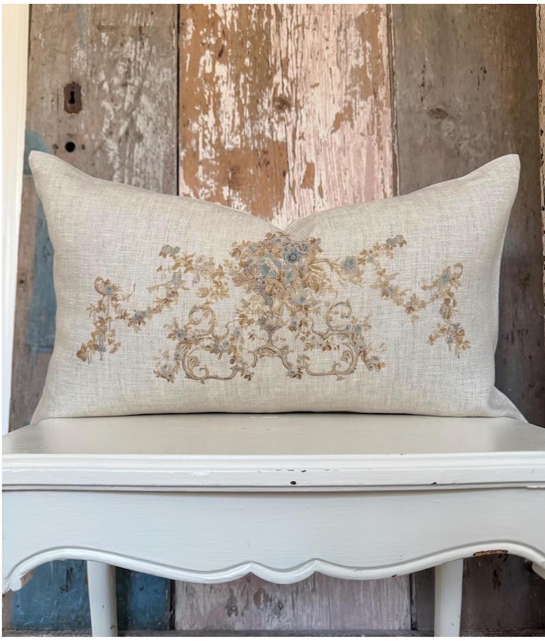 May include: A rectangular throw pillow made of light beige linen fabric. The center of the pillow features an intricate, symmetrical embroidered design in muted shades of dusty blue, sage green, and tan, depicting floral garlands and scrolls.