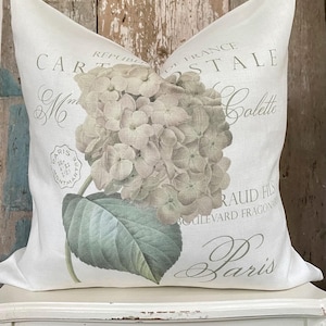 May include: White decorative pillow featuring a large, detailed hydrangea flower in muted green and gray tones. The pillow has French text and a vintage aesthetic, perfect for home decor.