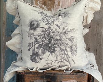 May include: A square, linen pillow with a floral print in shades of gray. The pillow has ruffled edges and is displayed on a small, dark wooden stand. The floral design features detailed botanical illustrations.