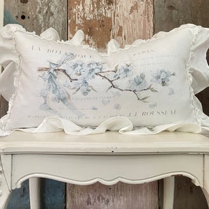May include: A white pillow with a floral design and the text "LA BOTANIQUE" and "PARIS" printed on it. The pillow has a ruffled edge and is sitting on a white table.